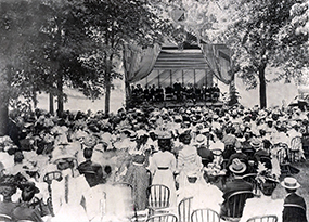 On a small stage with American flag drapery about 2 dozen young men dressed in suits are seated facing a large well-dressed crowd seated in chairs under trees.